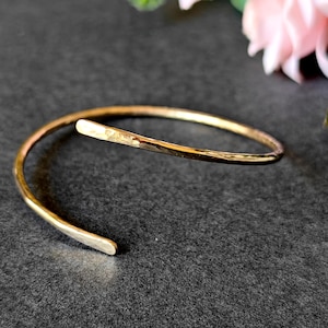 May include: A gold-colored, open cuff bracelet with a hammered texture. The bracelet has a simple, elegant design and is slightly curved. The ends of the bracelet are smooth and rounded. The bracelet is shown on a dark gray surface.
