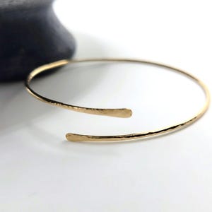 May include: A gold-colored open cuff bracelet with a hammered texture. The bracelet has an open design with slightly flattened ends. The jewelry is set against a white background with a dark gray object in the background.
