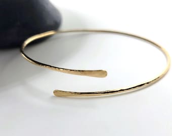 May include: A gold-colored open cuff bracelet with a hammered texture. The bracelet has an open design with slightly flattened ends. The jewelry is set against a white background with a dark gray object in the background.