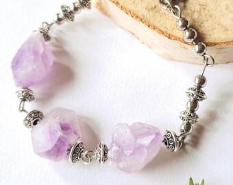 May include: A silver-toned bracelet featuring large, raw amethyst stones. The bracelet has a chain design with decorative silver beads and connectors. The amethyst stones are a light purple color and have a natural, irregular shape.
