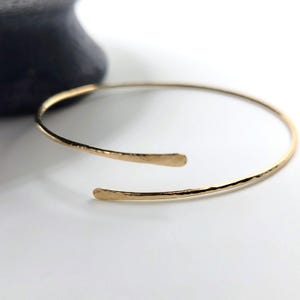 May include: A gold-coloured open cuff bracelet with a hammered texture. The bracelet has a simple, elegant design with slightly flattened ends. The jewellery is displayed on a white surface.