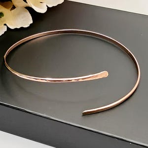 Puede incluir: Un brazalete de puño abierto en oro rosa con una textura martillada. El brazalete tiene un diseño simple y elegante y se muestra sobre una superficie negra. El diseño abierto permite un uso y ajuste fáciles. Una delicada pieza de joyería.