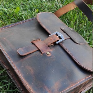 May include: A brown leather satchel bag with a buckle closure and a long strap. The bag has a distressed look and is sitting on a bed of green grass.