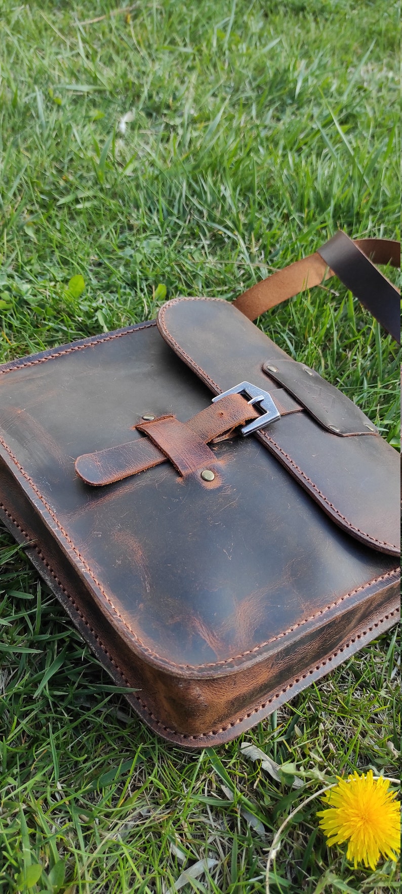 May include: A brown leather satchel bag with a buckle closure and a long strap. The bag has a distressed look and is sitting on a bed of green grass.