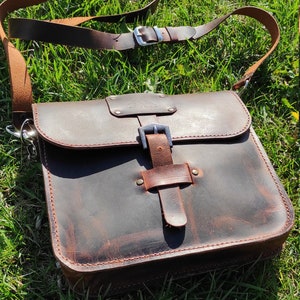 May include: A brown leather messenger bag with a buckle closure and a long adjustable strap. The bag has a distressed look and is made of high-quality leather.