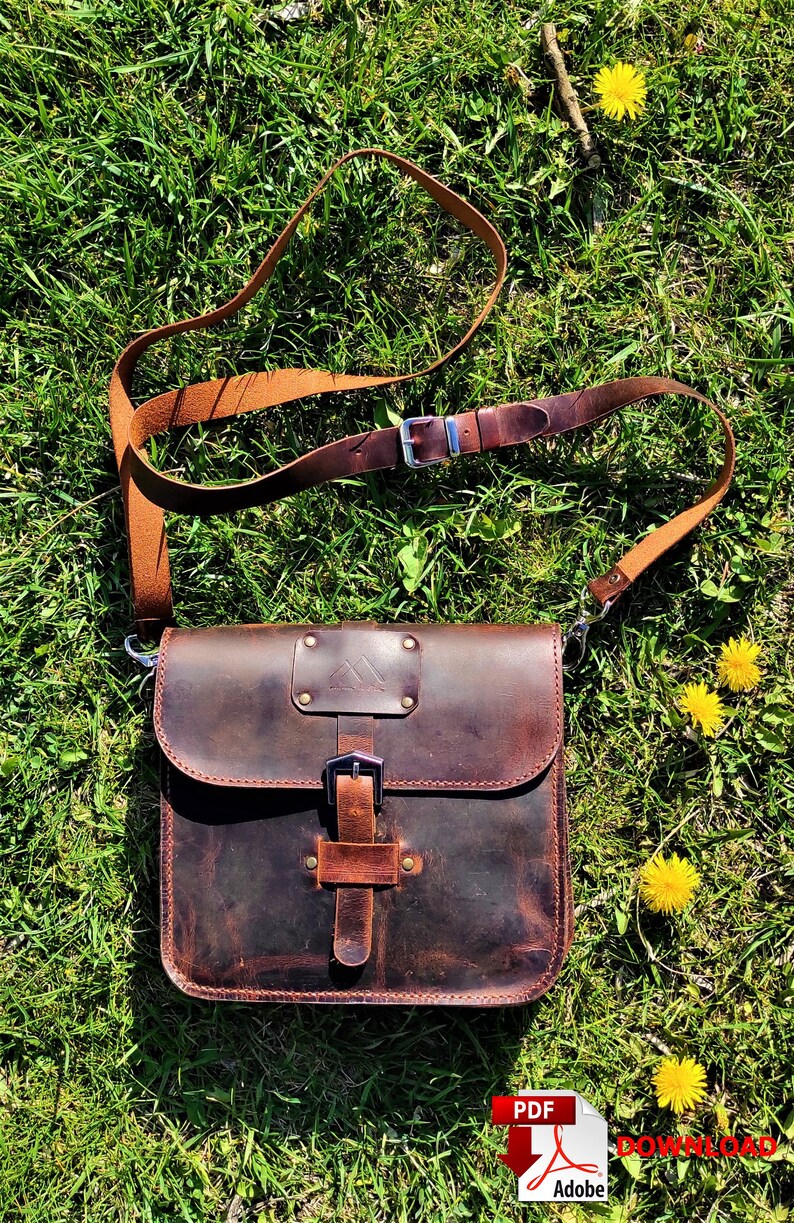 May include: A brown leather satchel bag with a long brown leather strap. The bag has a flap with a buckle closure and a small pocket on the front. The bag is sitting on a bed of green grass.