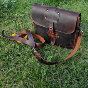 May include: A brown leather messenger bag with a buckle closure and a long adjustable strap. The bag has a distressed look and a small metal triangle logo on the front.