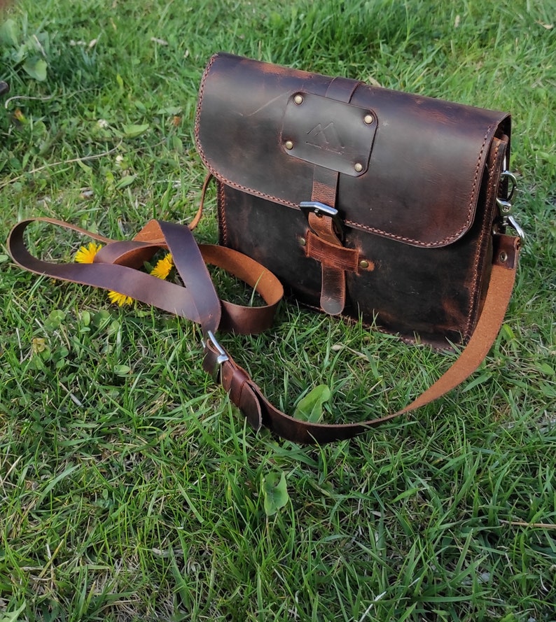 May include: A brown leather messenger bag with a buckle closure and a long adjustable strap. The bag has a distressed look and a small metal triangle logo on the front.