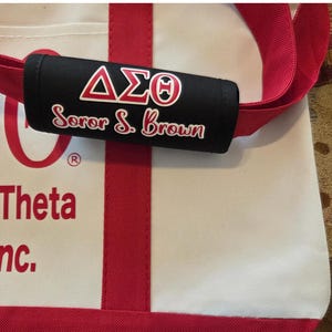 May include: A white tote bag with red accents and straps. The bag features a black handle wrap with the Greek letters &Delta;&Sigma;&Theta; and the words "Soror S. Brown" in white and red. The bag also has the word "Theta" in red.