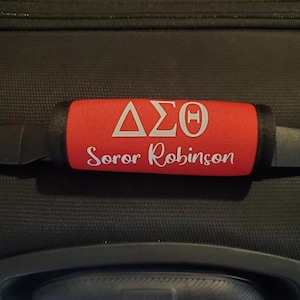May include: Red luggage handle wrap with white Greek letters and the name "Soror Robinson". The handle wrap is on a black suitcase. The letters are in a triangular and rectangular shape.