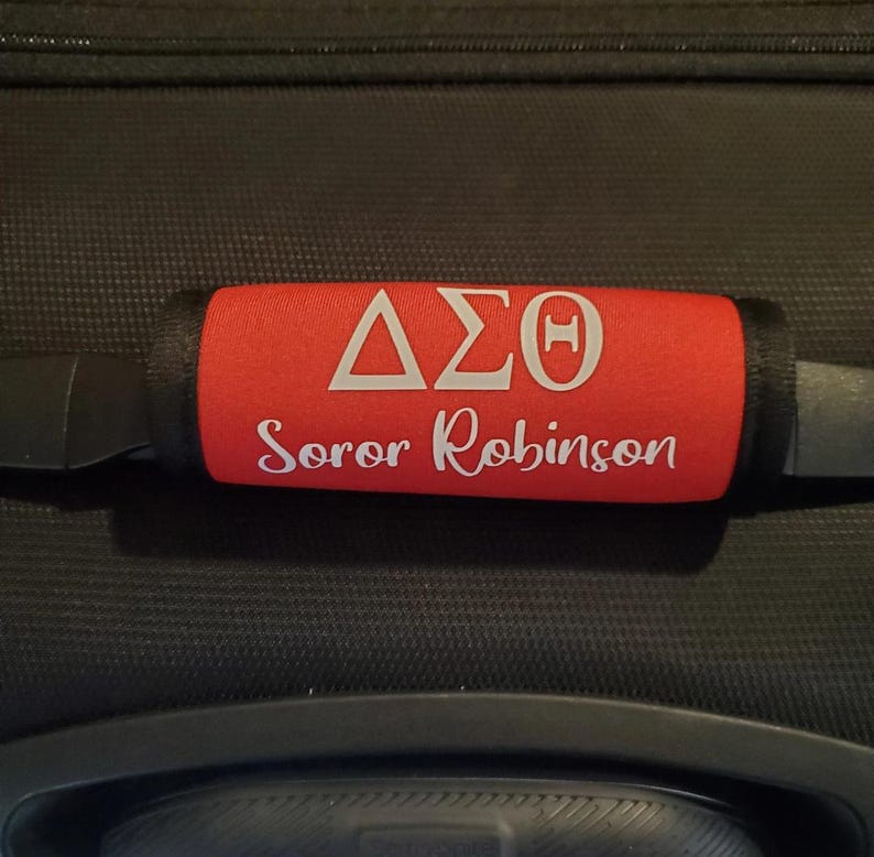 May include: Red luggage handle wrap with white Greek letters and the name "Soror Robinson". The handle wrap is on a black suitcase. The letters are in a triangular and rectangular shape.