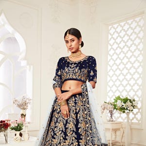 May include: A navy blue velvet lehenga with gold embroidery. The lehenga has a matching choli with a scoop neckline and short sleeves. The dupatta is a sheer white fabric with a gold border.