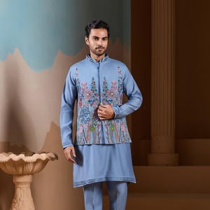 May include: A light blue ensemble featuring a long-sleeved tunic and matching trousers. The tunic is adorned with a floral embroidered vest. The outfit is completed with brown sandals. The background has a neutral color palette.