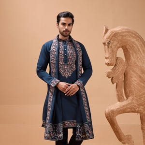 Puede incluir: Un hombre con un kurta azul marino con intrincados bordados florales en el escote y el chal. El chal y el kurta presentan un diseño de borde detallado. Lleva pantalones negros y sandalias marrones. Una escultura de caballo de madera está en el fondo.