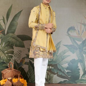 May include: A mustard yellow kurta and matching scarf with intricate embroidery, paired with white pants and brown sandals. The kurta features detailed patterns along the edges and sleeves. The outfit is worn by a person standing in front of a botanical mural.