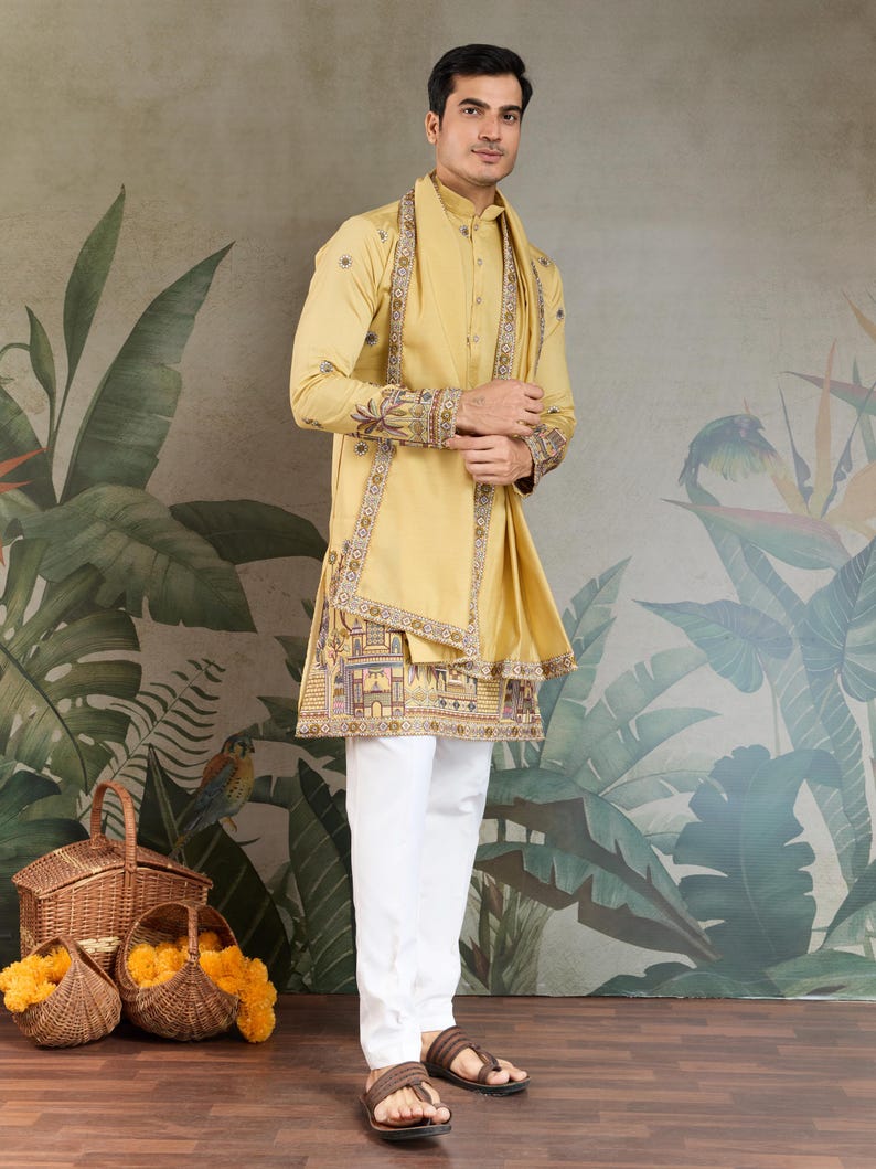 May include: A mustard yellow kurta and matching scarf with intricate embroidery, paired with white pants and brown sandals. The kurta features detailed patterns along the edges and sleeves. The outfit is worn by a person standing in front of a botanical mural.
