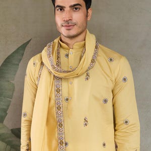 May include: A golden-yellow kurta pajama set with a matching scarf. The kurta features delicate floral embroidery and a mandarin collar. The scarf is draped around the neck and shoulders, with detailed patterned borders. The outfit is worn by a person.