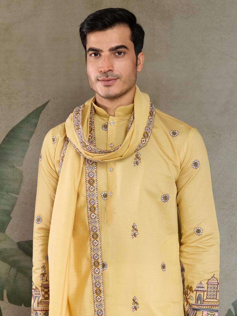 May include: A golden-yellow kurta pajama set with a matching scarf. The kurta features delicate floral embroidery and a mandarin collar. The scarf is draped around the neck and shoulders, with detailed patterned borders. The outfit is worn by a person.