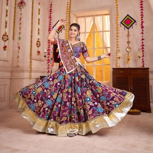 May include: A woman wearing a traditional Indian outfit. The ensemble features a purple skirt with floral patterns, a matching top, and a decorative scarf. The skirt has a gold trim. The outfit is adorned with colorful embroidery and embellishments.
