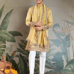 May include: A man wearing a mustard yellow kurta and matching scarf with intricate embroidery. The outfit is paired with white pants and brown sandals. The background features a botanical mural and baskets.