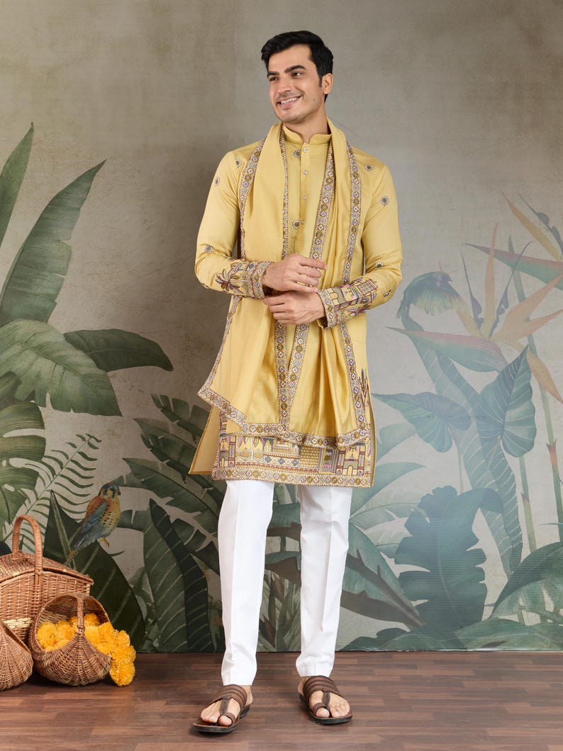 May include: A man wearing a mustard yellow kurta and matching scarf with intricate embroidery. The outfit is paired with white pants and brown sandals. The background features a botanical mural and baskets.