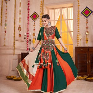 May include: A colorful traditional dress with a green, orange, and cream skirt. The bodice is green and orange with intricate embroidery. The dress features a decorative belt with tassels and is worn by a person in a room with decorative wall hangings.