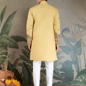 May include: A mustard yellow tunic with intricate embroidery on the sleeves, paired with white trousers. The tunic has a mandarin collar and a long, straight cut. The sleeves feature detailed patterns in shades of brown, red, and gold. The trousers are a crisp white, creating a contrasting look.