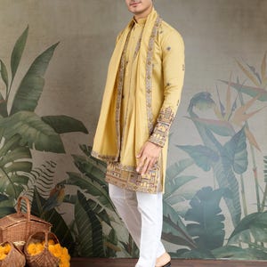 May include: A man wearing a mustard yellow kurta with intricate embroidery on the sleeves and hem, paired with white pants. A matching scarf is draped around the neck. The outfit is completed with brown sandals. The background features a floral mural.