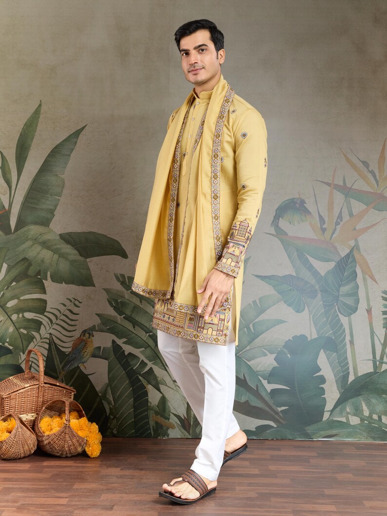 May include: A man wearing a mustard yellow kurta with intricate embroidery on the sleeves and hem, paired with white pants. A matching scarf is draped around the neck. The outfit is completed with brown sandals. The background features a floral mural.