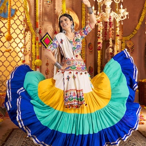 May include: A vibrant Indian dress featuring a full skirt in blue, turquoise, yellow, and white. The top is intricately embroidered with floral designs. The outfit is accessorized with silver jewellery.