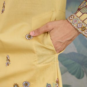 May include: Close-up of a mustard-yellow garment with floral embroidery. The garment features a pocket and is adorned with embroidered flowers in shades of blue, white, and brown. The sleeve cuff has intricate embroidery in similar colors.