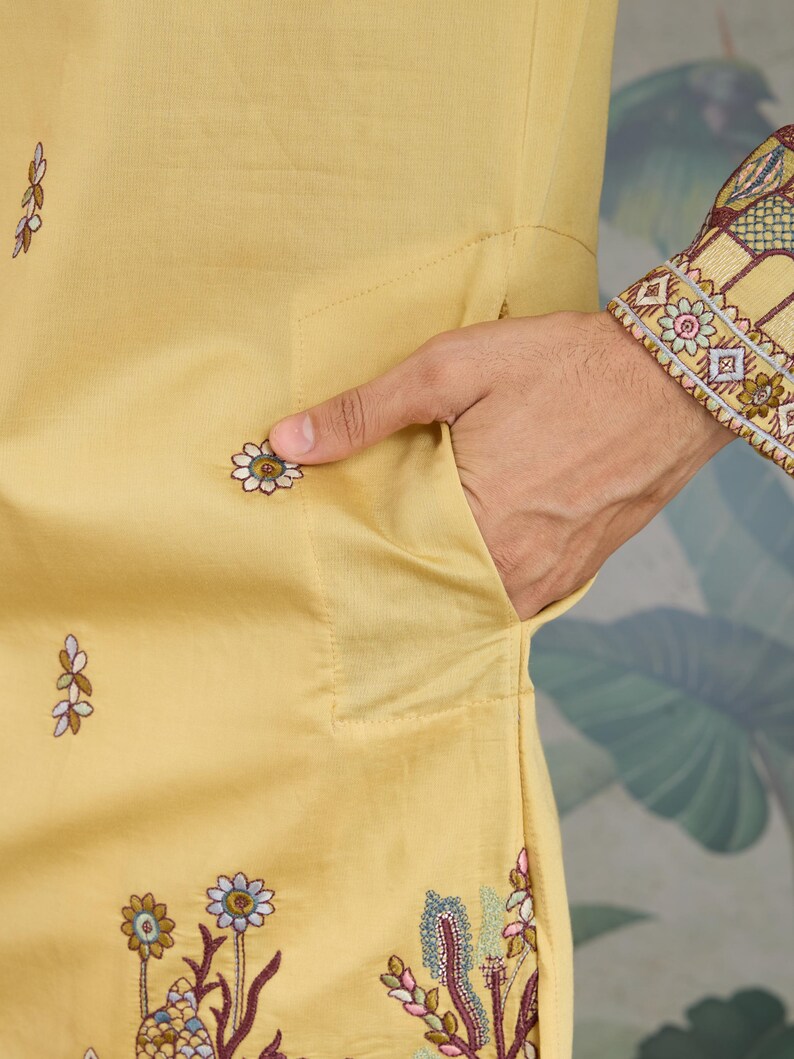 May include: Close-up of a mustard-yellow garment with floral embroidery. The garment features a pocket and is adorned with embroidered flowers in shades of blue, white, and brown. The sleeve cuff has intricate embroidery in similar colors.