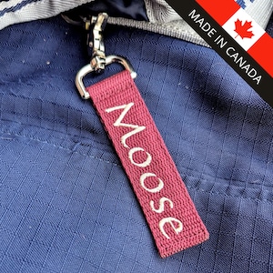 Peut inclure: Une étiquette en tissu bordeaux avec le mot "Moose" brodé en blanc, attachée à un fermoir en métal argenté. L'étiquette est contre un tissu bleu texturé. L'image comprend également une étiquette "Made in Canada".