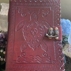 Puede incluir: Un diario de cuero burdeos con un diseño en relieve de tortuga marina en la portada. El diario tiene un cierre de metal y bordes cosidos. La cubierta es de un rico color marrón rojizo, de unos 15 cm de alto.