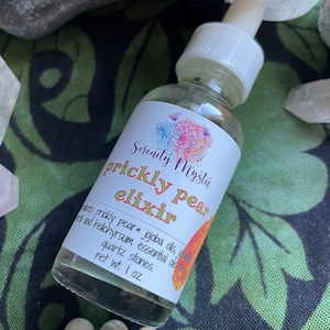 Puede incluir: Una pequeña botella de vidrio con un gotero blanco y una etiqueta que dice "Serenity Mystic prickly pear elixir". La etiqueta también incluye los ingredientes: pera espinosa, aceites de jojoba, jazmín y aceites esenciales de helicriso, piedras de cuarzo. La botella contiene un líquido transparente y está sobre una superficie verde con piedras blancas y verdes.