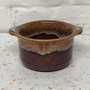 May include: A brown ceramic bowl with a dark brown exterior and a lighter brown interior. The bowl has two small handles and a textured rim. The bowl is on a white speckled surface.