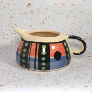 May include: A small, ceramic creamer with a cream-colored body and a black handle. The creamer is decorated with vertical stripes of orange, blue, and black, accented with green dots and a black circle. The spout is cream-colored.