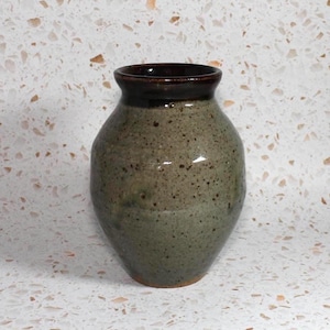 May include: A ceramic vase with a bulbous body and a flared rim. The main body features a speckled light green and grey glaze, while the rim is finished in a dark brown, glossy glaze. The base of the vase shows a hint of exposed terracotta clay.