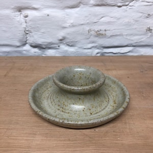 May include: A speckled, pale green ceramic candle holder. It features a small, raised cup in the centre, set on a circular base. The pottery has a rustic, handmade appearance, with visible texture and small brown speckles.