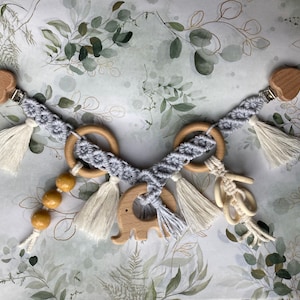 Könnte beinhalten: Ein graues und weißes gehäkeltes Babyspielzeug mit Holzringen, Perlen und einem Holzelefanten. Das Spielzeug hat Quasten und ist an zwei herzförmigen Holzklammern befestigt.