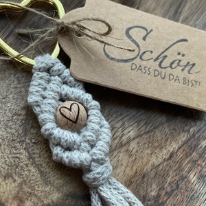 Könnte beinhalten: Ein grauer Makramee-Schlüsselanhänger mit einer Holzperle, die ein Herzdesign aufweist. Der Schlüsselanhänger hat einen goldenen herzförmigen Ring und ein braunes Etikett mit dem Text "Schön, dass du da bist!"