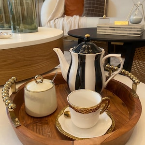 May include: A wooden serving tray holds a black and white striped teapot, a sugar bowl, and a teacup with a gold rim. The tray has gold handles and a gold rim on the teacup and saucer. The tea set is on a white surface.
