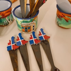 May include: Wooden spoons with a dark brown finish and a painted Dominican Republic flag design on the handle. The spoons are displayed in a small metal cup with a painted landscape design. Other cups with similar designs are also visible.