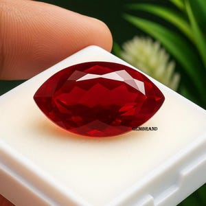 May include: Marquise-cut, faceted, deep red gemstone. The gem is displayed on a white surface, with the text "GEMBRAND" visible. The gemstone's facets reflect light, showcasing its brilliance. The overall presentation suggests a focus on jewelry or gemstones.
