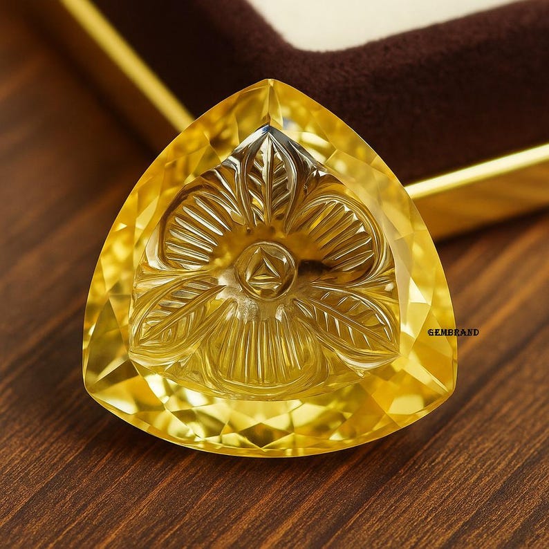 Puede incluir: Una gema triangular facetada con un dise&ntilde;o floral tallado. La gema es de color amarillo vibrante y parece estar colocada en una caja. La palabra "GEMBRAND" es visible en la parte inferior derecha de la imagen.