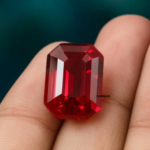 May include: A faceted, rectangular-cut, deep red gemstone. The gem has a high degree of clarity and brilliance, reflecting light from multiple facets. The gemstone is held in a hand, with the Gembrand logo visible.