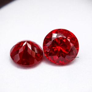 May include: Two faceted, round-cut, deep red gemstones. The gems are highly reflective, with multiple facets catching the light. The gems are set against a white background, highlighting their vibrant color and clarity. The gemstones are likely rubies.