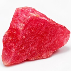 May include: A rough-cut, vibrant red gemstone with a textured surface. The stone has a natural, irregular shape and a slightly translucent appearance. The color is a deep, saturated red, with variations in tone across the surface.
