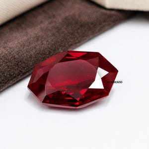 Könnte beinhalten: Ein facettierter, tiefroter Edelstein mit Brillantschliff. Der Edelstein liegt vor einem weißen Hintergrund mit einem braunen Stoff in der oberen linken Ecke. Der Text "GEMBRAND" ist auf der rechten Seite des Edelsteins zu sehen.