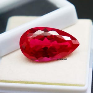 Puede incluir: Una gema roja vibrante en forma de pera, con un corte facetado, se muestra sobre un fondo blanco. La gema está en una pequeña caja con la palabra "GEMBRAND" impresa en la parte inferior.